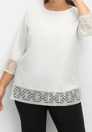 Mujer con blusa blanca con adornos de encaje floral en las mangas y el dobladillo, combinada con pantalones negros, sobre un fondo liso.