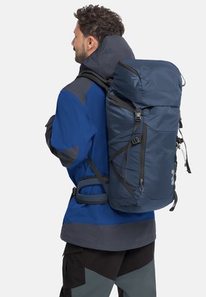 Homme portant une veste bleue et grise, portant un grand sac à dos de randonnée bleu marine avec des sangles, sur fond blanc.
