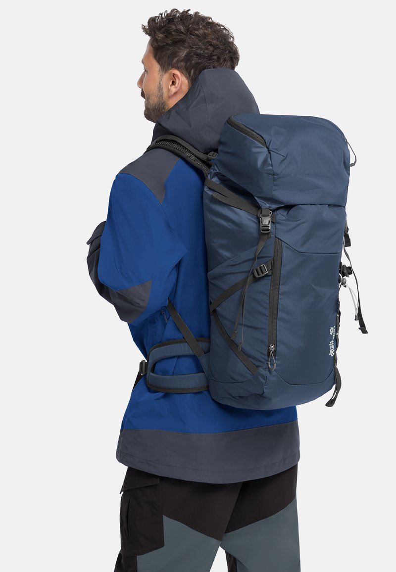 Homme portant une veste bleue et grise, portant un grand sac à dos de randonnée bleu marine avec des sangles, sur fond blanc.