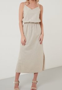 Beige maxi šaty bez rukávů s V-výstřihem, elastickým páskem, bočním rozparkem a měkkou, texturovanou látkou. V kombinaci s světlými sandály na kotníky.