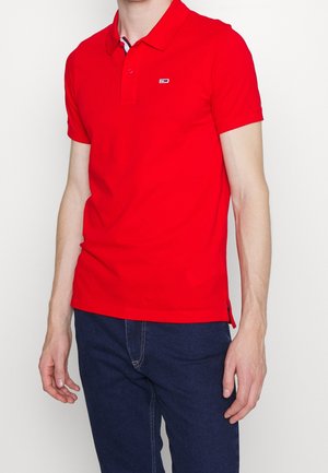 Czerwona koszulka polo wykonana z bawełny, z kołnierzykiem, krótkim rękawem i małym logo na piersi. W zestawie z ciemnoniebieskimi dżinsami.