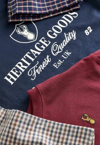 Tre klädesplagg: en marinblå sweatshirt med vit text och älglogo, en vinröd stickad tröja med broderad detalj, och ett rutig tyg.