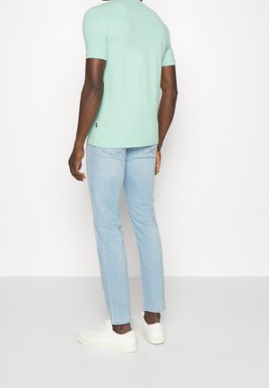 Hombre con jeans azul claro, zapatillas blancas y una camiseta tipo polo de manga corta verde menta, de pie contra un fondo blanco, vista desde atrás.