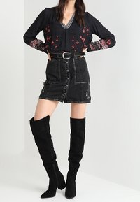 Svart blommig blus med lång ärm, kombinerad med en svart denimkjol med knappar och sidobroderi, kompletterad med knähöga svarta stövlar.