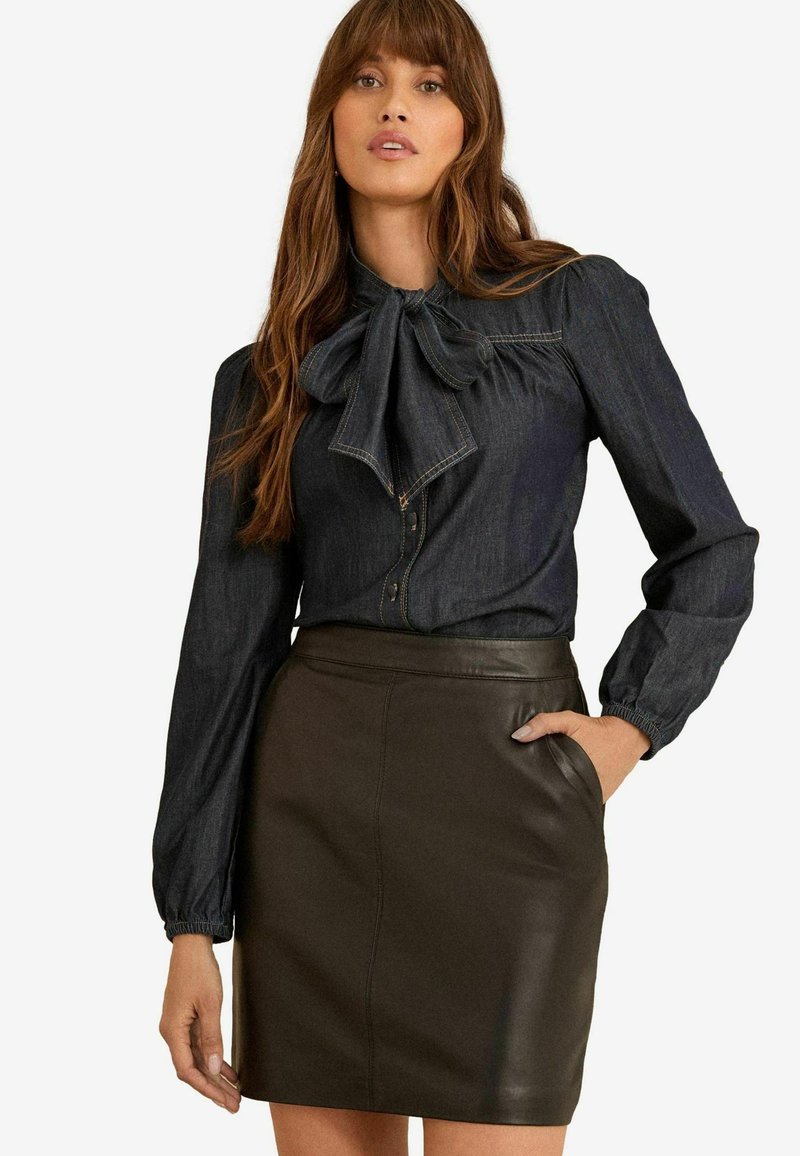 Blusa di jeans scuro con colletto a fiocco, maniche a palloncino e chiusura a bottone, abbinata a una gonna in pelle marrone scuro aderente.