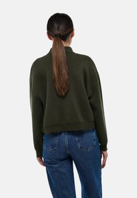 Pull en maille vert foncé à coupe décontractée, avec des poignets côtelés et un ourlet court, associé à un jean taille haute bleu moyen.