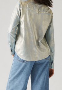 Camicia blu metallizzata con orlo curvo e polsini abbottonati, abbinata a jeans blu chiaro dal taglio morbido. È visibile una leggera lucentezza.