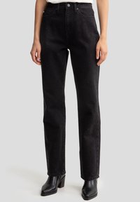Vaqueros de mezclilla negra con un diseño de pierna recta, que cuentan con sutiles adornos y una cintura alta. Llevados con botas de tacón negro hasta el tobillo.
