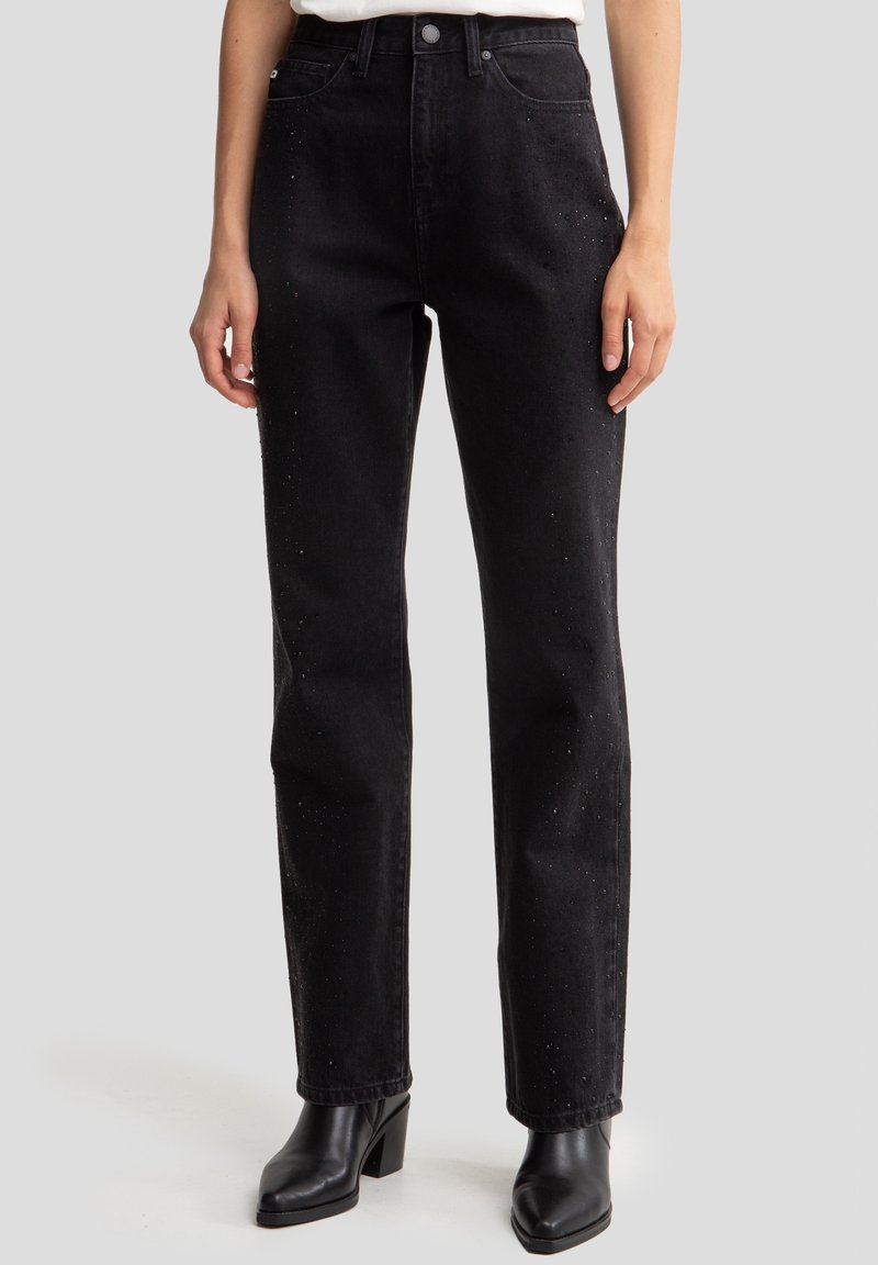 Vaqueros de mezclilla negra con un diseño de pierna recta, que cuentan con sutiles adornos y una cintura alta. Llevados con botas de tacón negro hasta el tobillo.