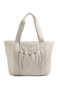 Sac fourre-tout en tissu beige avec de larges poignées et une poche à élastique sur le devant, texture douce, forme rectangulaire.