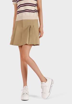 Gonna plissettata color khaki abbinata a un top in maglia a righe; il modello indossa sneakers bianche con una texture liscia e accenti neri a contrasto.