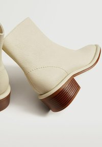 Bottes en cuir beige à la cheville avec une texture lisse, un bout arrondi et un talon en bois nervuré. Des coutures simples viennent agrémenter le design.