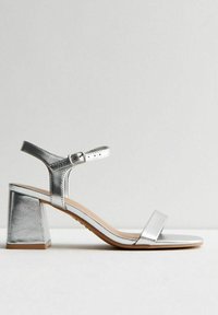 Silver metallisk klacksandaler med blockklack, ankelsnodd och minimalistisk design; har platt sula och en enda framrem.