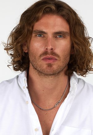 Uomo con capelli ondulati, lunghi fino alle spalle e una leggera barba, che indossa una camicia bianca con bottoni e una collana di argento con un pendente unico.