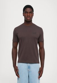 Tricou scurt cu mânecă brown dintr-un material moale, având un gât rotund și un mic logo negru EA7 pe pieptul stâng. Asortat cu pantaloni din denim deschis.