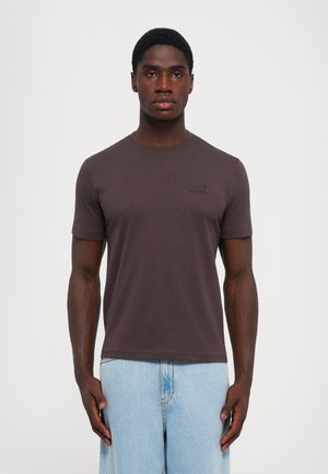 Tricou scurt cu mânecă brown dintr-un material moale, având un gât rotund și un mic logo negru EA7 pe pieptul stâng. Asortat cu pantaloni din denim deschis.