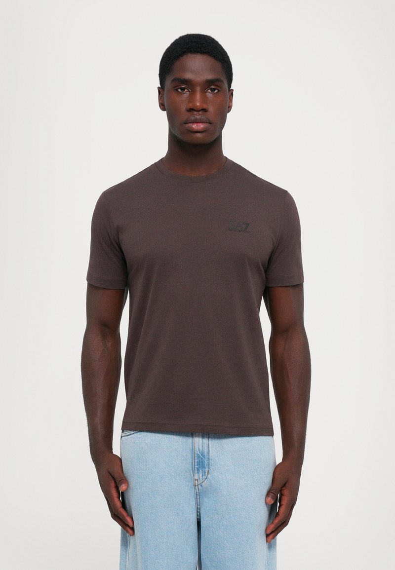 Tricou scurt cu mânecă brown dintr-un material moale, având un gât rotund și un mic logo negru EA7 pe pieptul stâng. Asortat cu pantaloni din denim deschis.