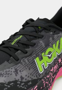 Gros plan d'une chaussure de course noire et grise avec le logo "HOKA" vert néon, des lacets noirs et un détail de semelle tacheté rose.