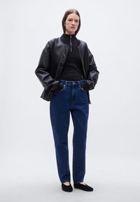 Μαύρο μπουφάν bomber με καπιτονέ ύφασμα πάνω από μαύρο πουλόβερ ζιβάγκο, σε συνδυασμό με τζιν παντελόνι ίσιας γραμμής σκούρου μπλε και μαύρα παπούτσια χωρίς κορδόνια.