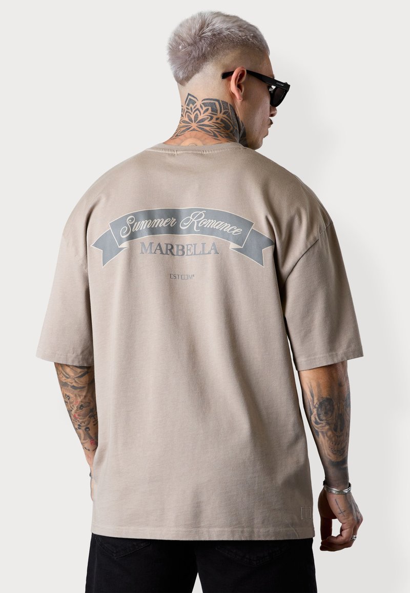 T-shirt beige surdimensionné avec un design graphique gris au dos. Présente le texte "Summer Romance" et "MARBELLA". Matière en coton doux.