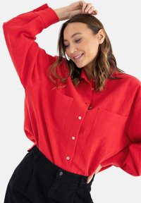 Czerwona, oversize koszula z guzikami, wykonana z fakturowanej tkaniny, z wyraźnym kołnierzykiem i dwiema kieszeniami z przodu, zestawiona z czarnymi spodniami.