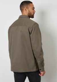 Olijfgroene canvas jas met een kraag, lange mouwen en knoopmanchetten. Heeft een yoke-naad aan de achterkant en subtiele stikdetails.