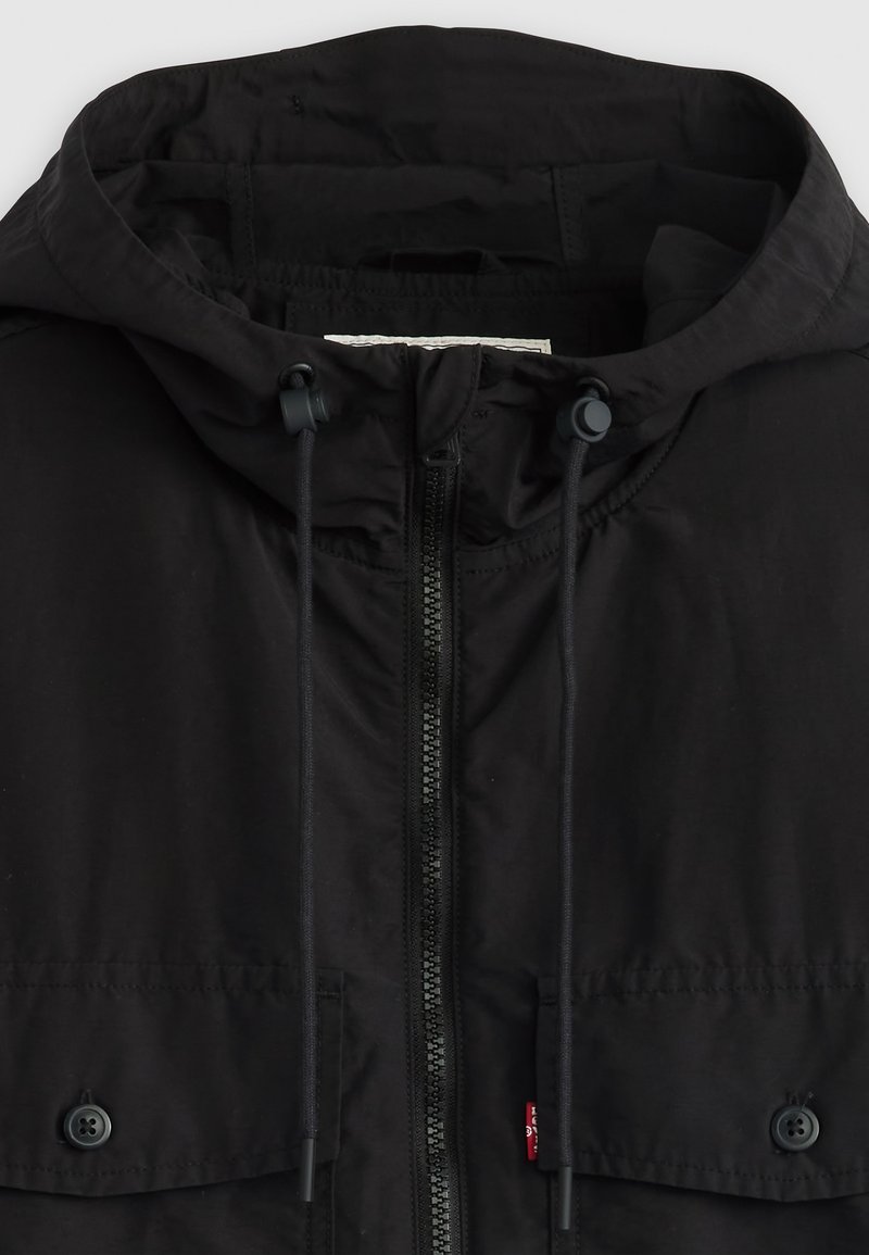 Chaqueta con capucha negra hecha de tejido ligero, que presenta un cierre completo en la parte delantera, cordones ajustables y dos bolsillos con botón.