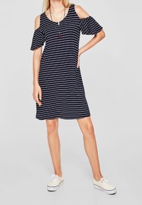 Kvinna iklädd en marinblå och vit randig klänning med kalla axlar och korta volangärmar, stående med en hand på höften.