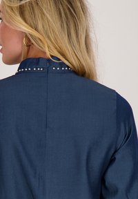 Marineblauwe blouse van gestructureerde stof, met een hoge kraag en decoratieve zilveren studs langs de halslijn aan de achterkant.