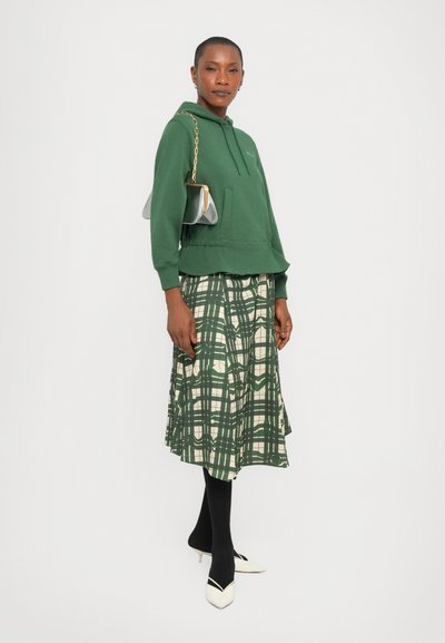 Felpa verde con tasca frontale e cordoncino, abbinata a una gonna a quadri verde e crema. Accessoriata con una borsa a tracolla argento.