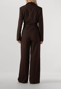 Bruine op maat gemaakte jumpsuit met lange mouwen, gestructureerde schouders en een riem in de taille. Bevat brede broekspijpen en een gladde stoftextuur.