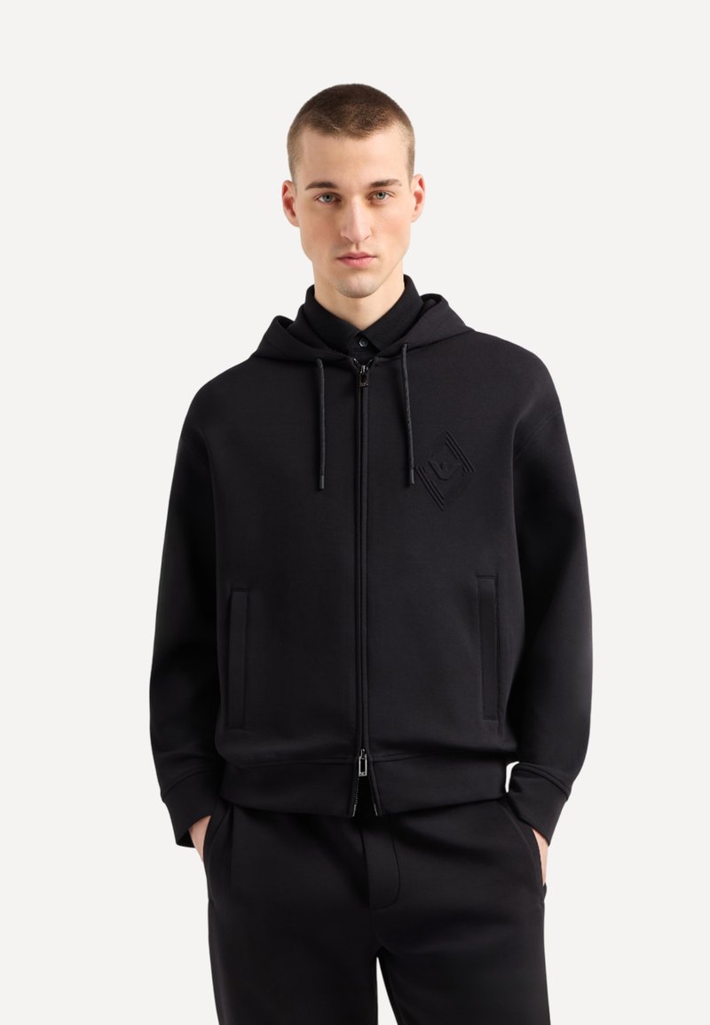 Hoodie noir zippé en tissu doux, avec une capuche, des poches avant et un léger détail de logo embossé. Texture lisse et coupe décontractée.