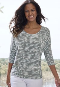 Hellblau-grünes Langarmoberteil mit einer wellenförmigen weißen Musterung, Rundhalsausschnitt und lockerer Passform, kombiniert mit weißen Hosen.