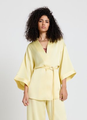 Frau mit lockigem Haar, die ein blassgelbes Kimono-Oberteil mit weiten Ärmeln und passende Hosen trägt, accessoriert mit goldenen Ringen und einer Halskette.