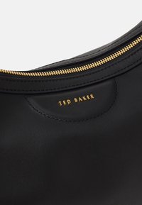 Černá kožená taška se zlatým zipem a vyraženým logo "TED BAKER". Hladký povrch a zaoblený design s viditelnými švy.