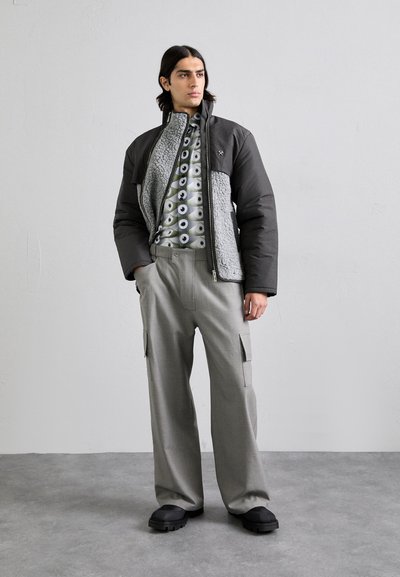 Dunkelgraue Pufferjacke mit einem strukturierten grauen Fleece-Innenfutter, kombiniert mit einem gemusterten Hemd mit kreisförmigen Designs und lockeren grauen Hosen.