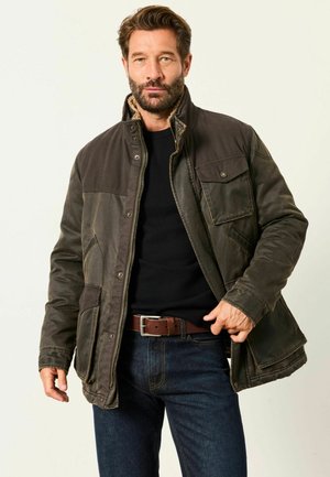 Mann trägt dunkelgrüne Jacke, schwarzen Pullover, braunen Gürtel und blaue Jeans, zieht den Reißverschluss der Jacke hoch und blickt nach vorne.