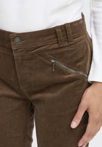 Rudi korduroviniai kelnės su vertikaliais briauneliais, priekyje su užtrauktuku uždaryta kišene ir su sagos užsegimu liemens srityje.