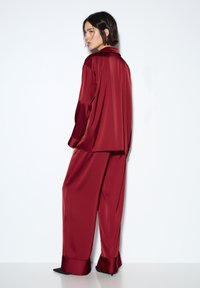 Dvoubarevný saténový komplet ve vínové barvě, sestávající z volně střižené košile na knoflíky a kalhot s širokými nohavicemi zakončenými manžetami. Hladká textura, elegantní splývavost.