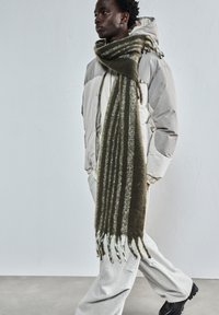 Mlad oseba oblečena v bež puhovko, dolgo črtasto zeleno šalo, svetlo siva hlače in črne čevlje, hodi ob enobarvni svetlo sivi steni.