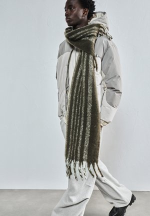 Fiatal személy bézs színű puffertakót, hosszú csíkos zöld sálat, világosszürke nadrágot és fekete cipőt viselve, egy sima világosszürke fal előtt sétál.