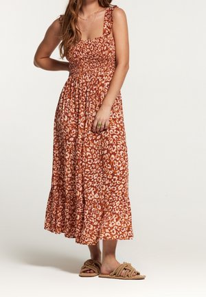 Mujer con vestido midi sin mangas con estampado en tonos óxido y crema, con tirantes con volantes y sandalias planas estilo cuerda, de pie con una mano en la cadera.