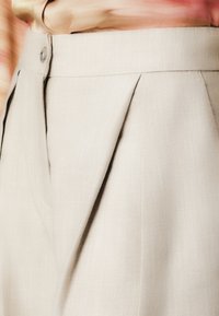 Μπεζ πλισέ παντελόνι κατασκευασμένο από υφασμάτινη υφή, με κουμπί στο φερμουάρ και εφαρμογή κομμένη με καθαρές γραμμές.