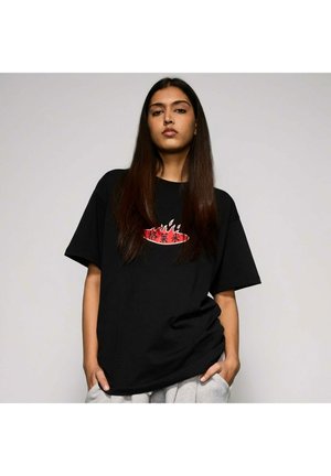 Mujer joven con cabello largo y oscuro, lleva una camiseta negra con un diseño de llamas rojas y blancas y caracteres asiáticos, con las manos en los bolsillos de unos pantalones grises.