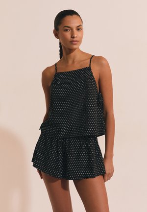 Kobieta z warkoczami, ubrana w czarną, bezrękawnikową bluzkę w groszki i pasujące krótkie spodenki, stojąca na jednolitym beżowym tle.