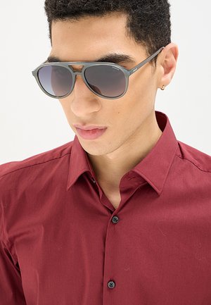 Uomo con occhiali da sole scuri e una camicia rossa abbottonata con un piccolo orecchino a cerchio d’oro nell’orecchio sinistro.