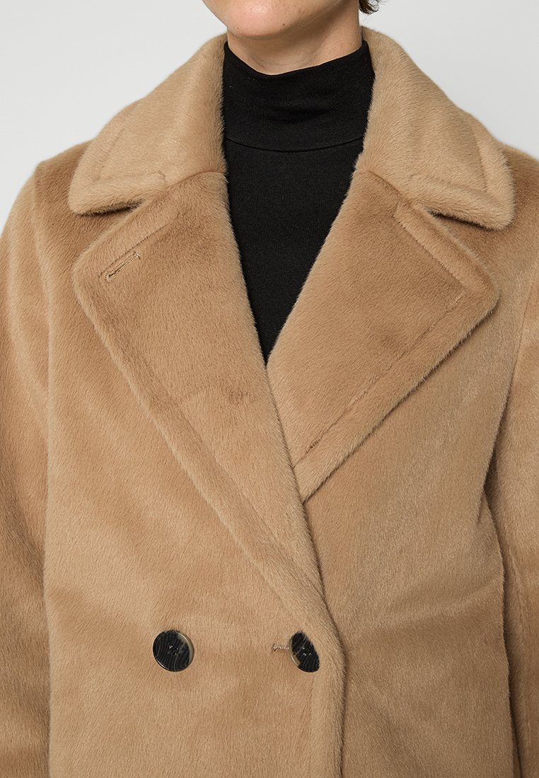 Beżowy płaszcz podwójnie zapinany, wykonany z miękkiego materiału przypominającego futro, z dużymi czarnymi guzikami i klapami. Teksturowana powierzchnia dodaje głębi.