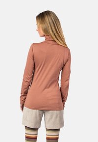 Langärmliges Rollkragenoberteil in Terrakottafarbe, aus leichtem Stoff gefertigt. Kombiniert mit beigen Cordshorts und gestreiften Beinstulpen.