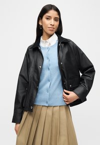 Γυναίκα που φοράει μαύρο μπουφάν πάνω από ανοιχτόχρωμο μπλε πουλόβερ με κουμπιά και λευκό πουκάμισο με γιακά, συνδυασμένο με μπεζ πλισέ φούστα, στέκεται μπροστά σε λευκό φόντο.