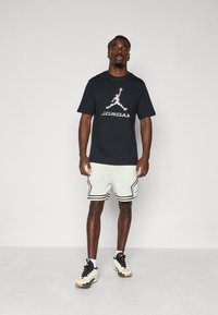 Sort t-shirt med hvid Jordan-logo, lysfarvede perforerede shorts med sort kant og sneakers med en blanding af hvid og sort.